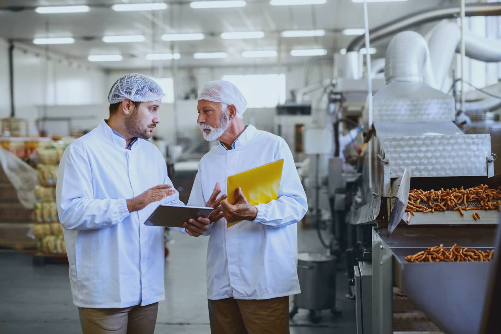 Two men in a food processing plant discussing operations next to a machine producing french fries