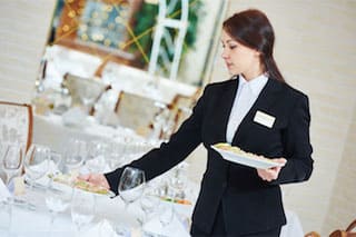 Female catering staff member wearing suit working at an event at an upscale venue. She is setting food presented on fine china on a table.