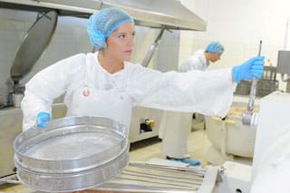 Uniformed female food processing plant worker turning lever on machinery while holding large metal cake pan