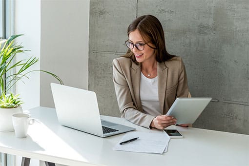 Businesswoman in blazer holding a tablet and smiling while looking at her laptop screen in a sunny, modern office
