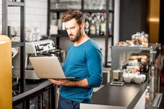 Restaurant manager dressed in jeans and long sleeve shirt holding a laptop and glancing at the screen from behind the cafe counter