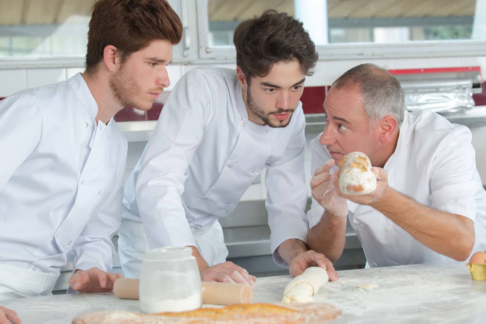 A senior chef holding bread up as two younger chefs examine the composition