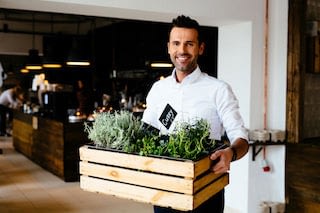 Happy restaurant manager carrying a crate of fresh herbs outside seating area of restaurant
