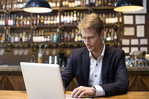 Male restaurant manager in blazer reviewing data dashboards on laptop at restaurant bar